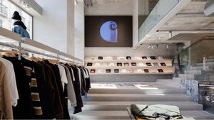 The image shows a modern clothing store interior with racks of hanging clothes on the left and neatly folded shirts on shelves in the background. A large abstract artwork is displayed on the wall above the shelves, and the space features concrete and glass elements.