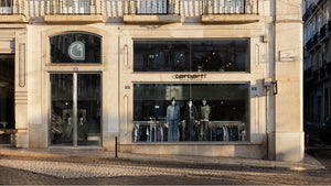 The image shows the exterior of a Carhartt WIP store with mannequins displaying clothing in the window, set in a historic building with a cobblestone street in front.