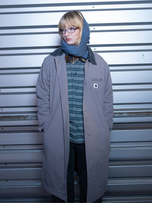 A person wearing glasses and a scarf stands against a metal wall, wearing a coat with a visible Carhartt WIP logo.