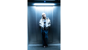 A person stands in front of an elevator wearing a light-colored jacket and dark pants, possibly from Carhartt WIP.