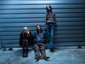 Three people in casual attire sit and stand against a metal wall, with no visible Carhartt WIP branding.